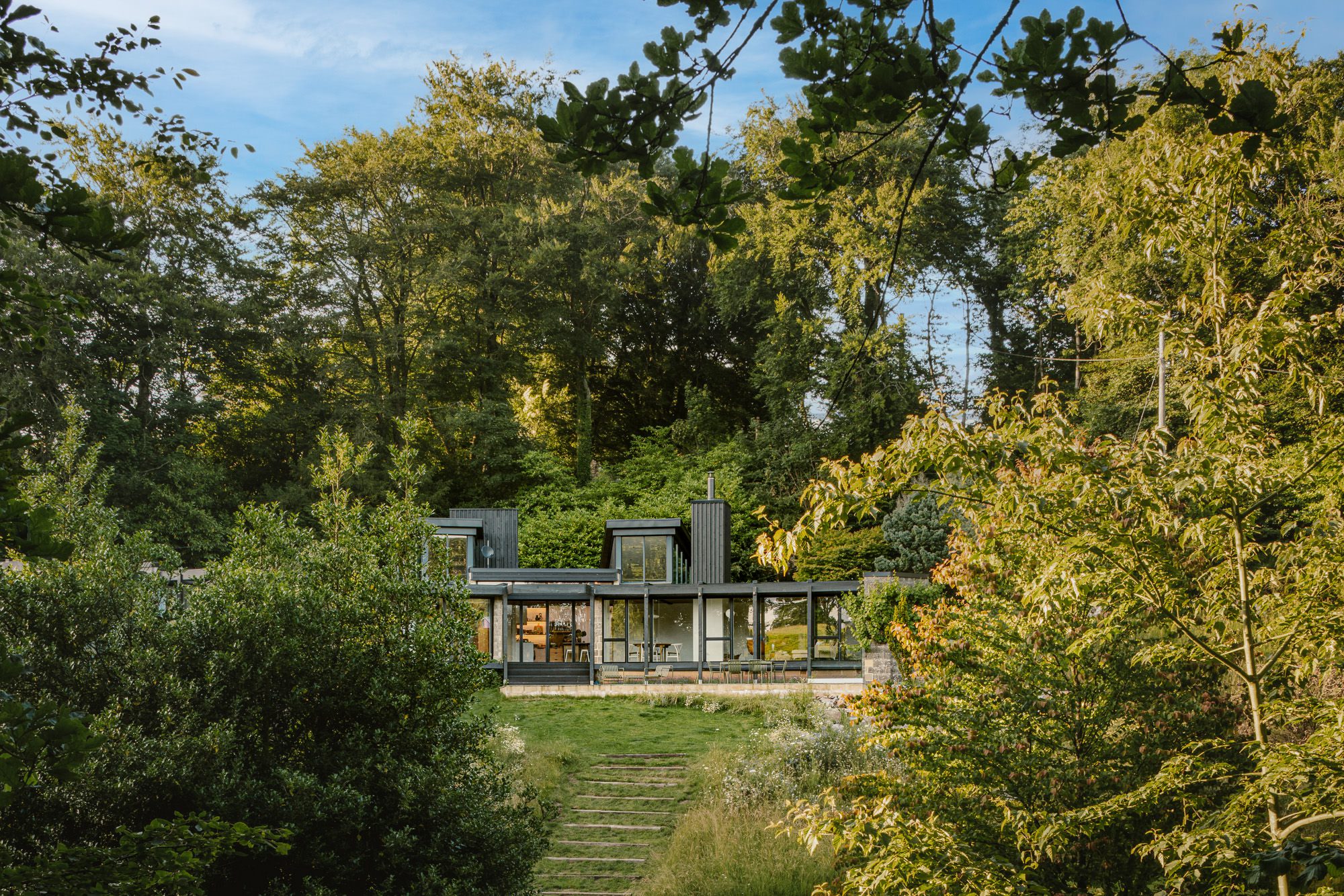 Capture from the bottom, featuring the upper portion of a house surrounded by trees with a clear sky in the background, and showcasing a window of the home, photographed by Brett Charles for building photography in the UK