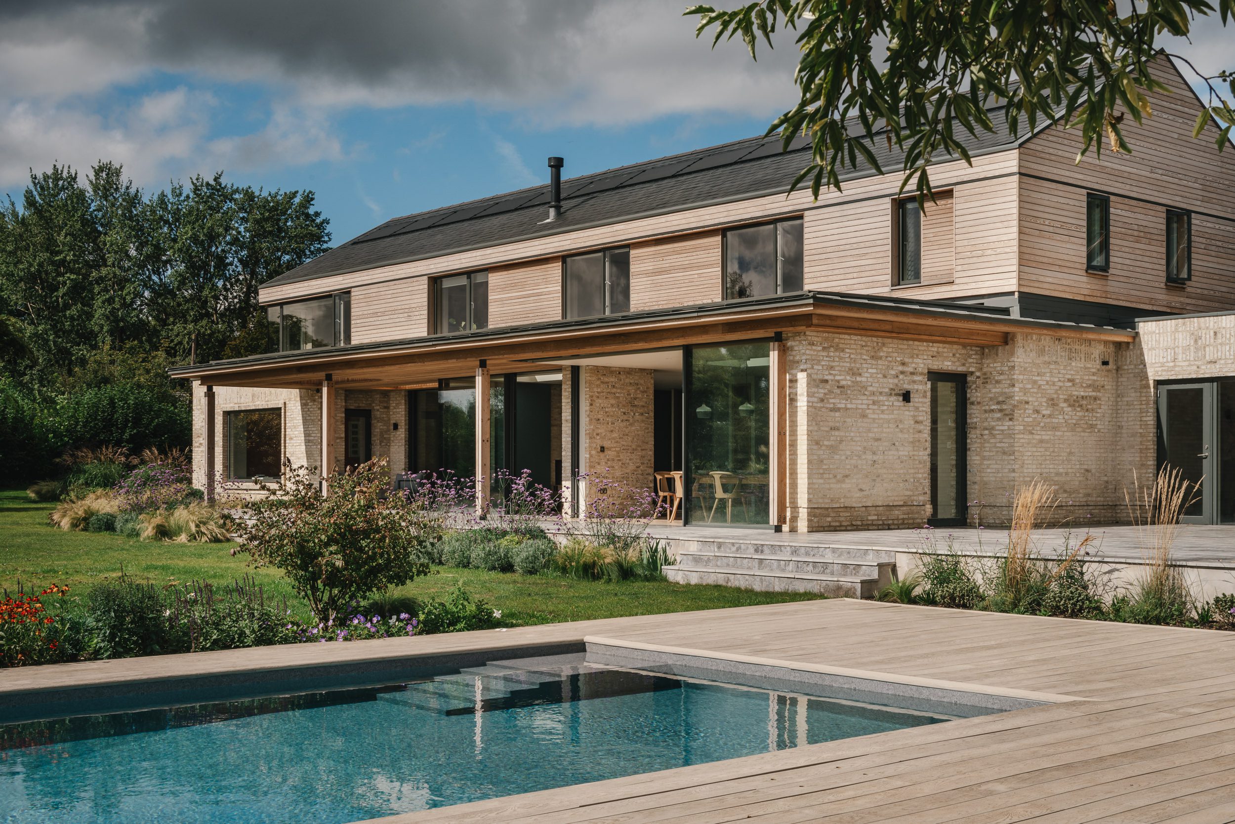 Side view of a house captured from the other side of a swimming pool within the house, with green lush grass in front, photographed by Brett Charles, showcasing architectural photography in the UK.